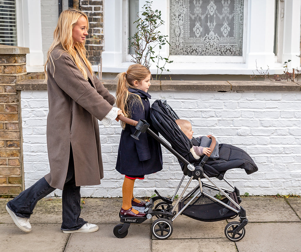 A woman pushes a black stroller with a baby inside down an pavement in front of a white brick house. A young girl stands on the attached Lascal buggy board.