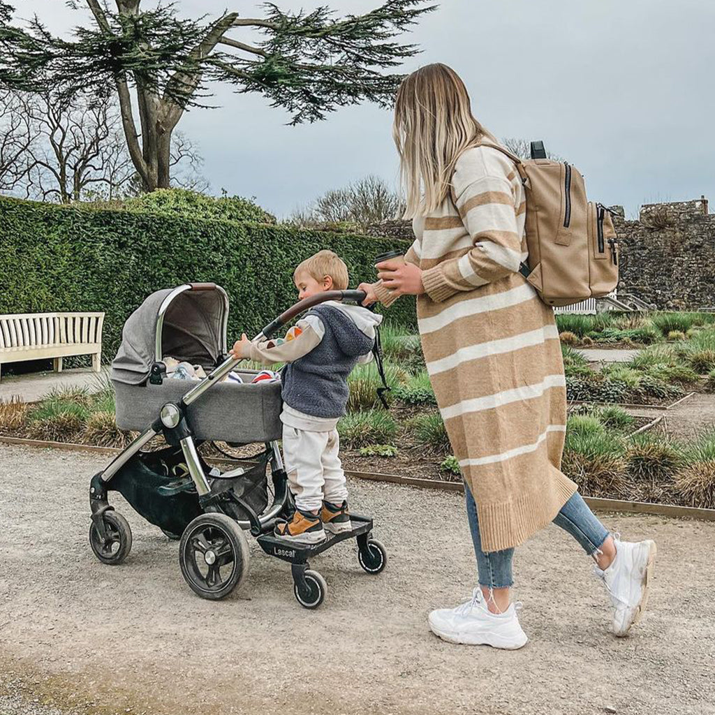 A woman in a beige and white striped knit sweater and blue jeans walks beside a stroller with a young child standing on a Lascal buggy board, outdoors on a gravel path with hedges and trees in the background.